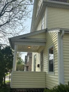 Side view of a pale yellow house with white trim and a covered front porch, with bare tree branches in the foreground.