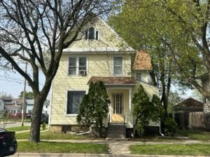 Front view of a pale yellow two-story house with a pitched roof and dormer window, framed by leafing trees on a sunny day with a small front porch.