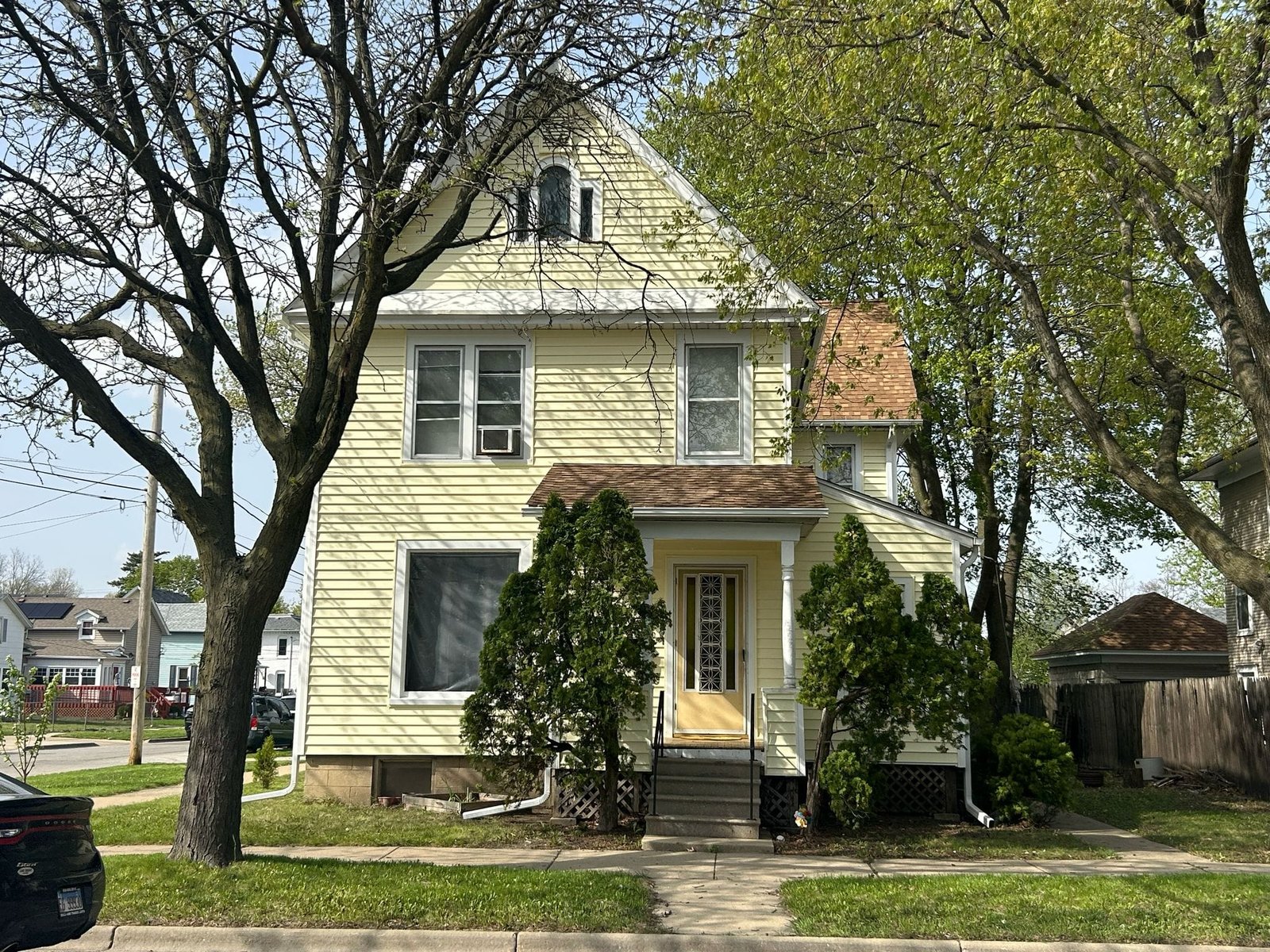 Front view of a pale yellow two-story house with a pitched roof and dormer window, framed by leafing trees on a sunny day with a small front porch.