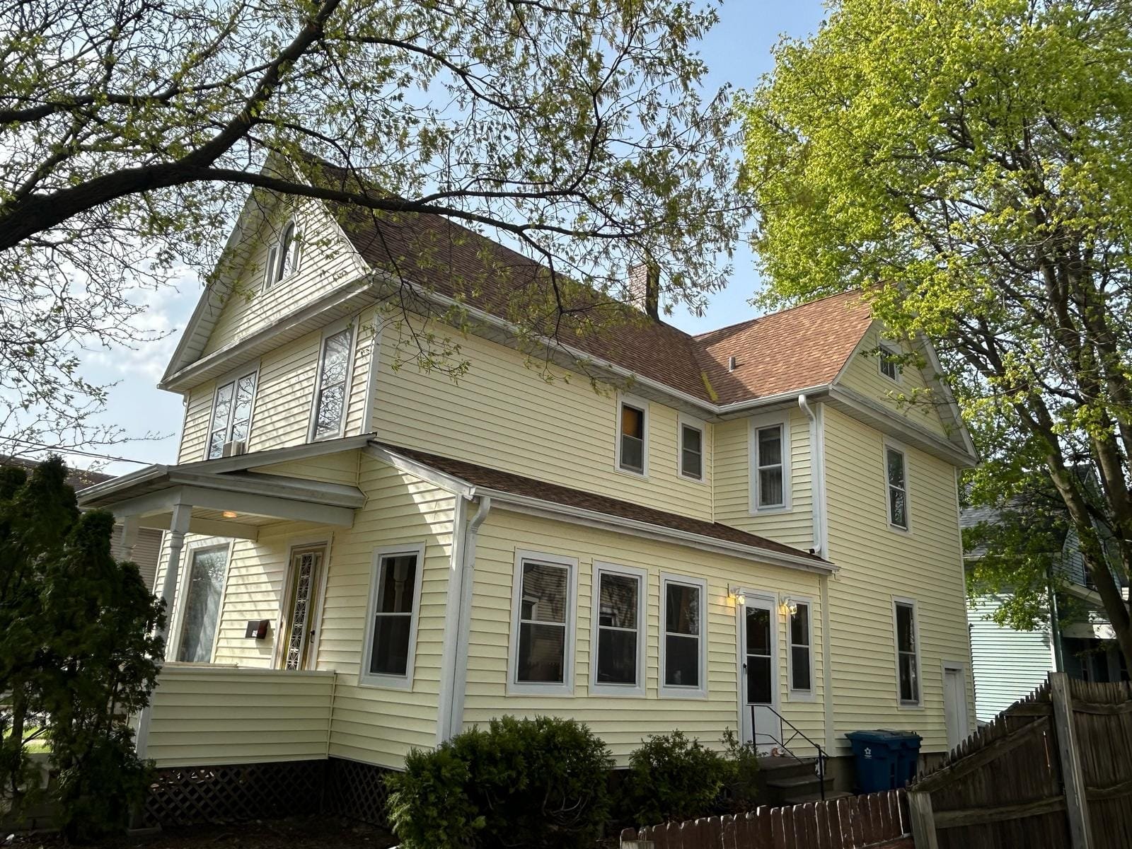 Yellow two-story house with a steep gabled roof, white trim, and a small covered porch, set among leafy trees on a sunny day.