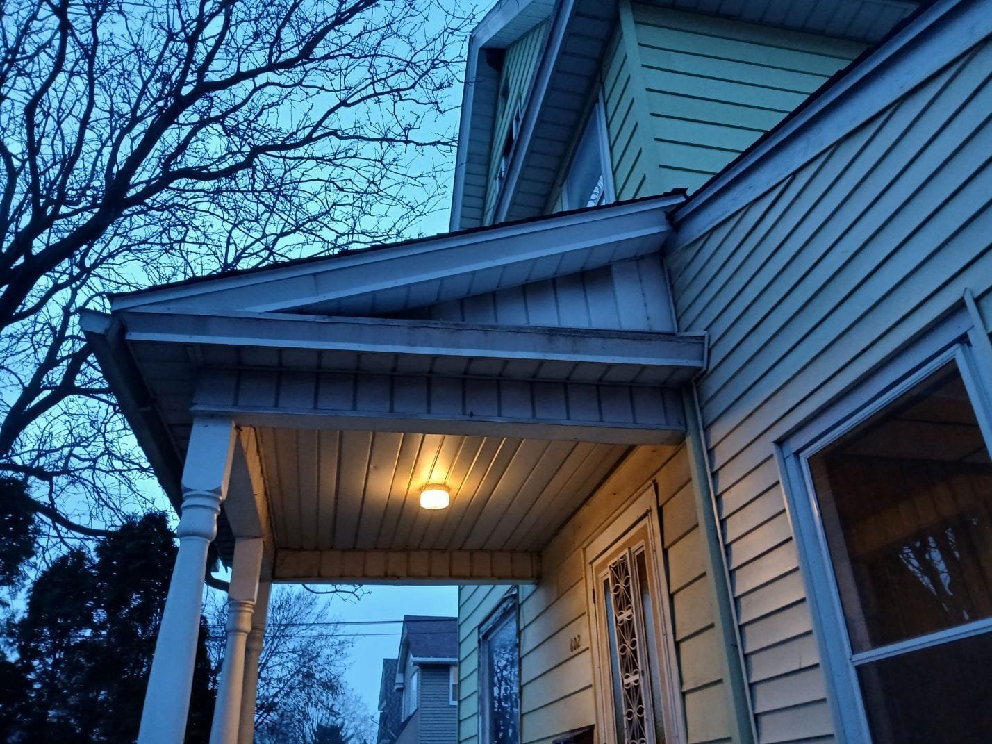 Front porch of a yellow house at dusk with white columns, a lit porch light, and a decorative door grille.