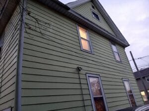 Green two-story house with horizontal siding and multiple rectangular windows; a security camera is mounted on the wall and power lines cross the scene at dusk.