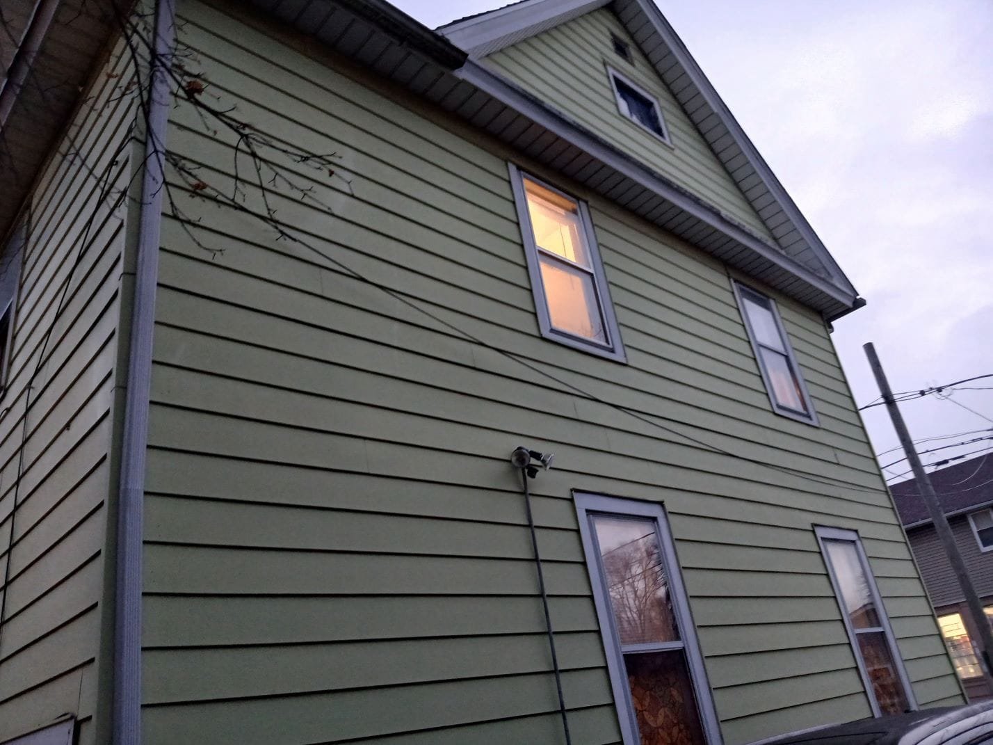 Green two-story house with horizontal siding and multiple rectangular windows; a security camera is mounted on the wall and power lines cross the scene at dusk.