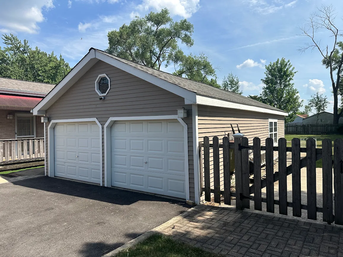 Two-car detached garage with beige siding, white raised-panel doors, and an octagonal window under a gabled roof.