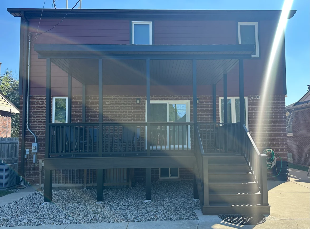 Two-story brick house with a dark wooden elevated deck and covered porch, accessed by stairs, overlooking a gravel yard.