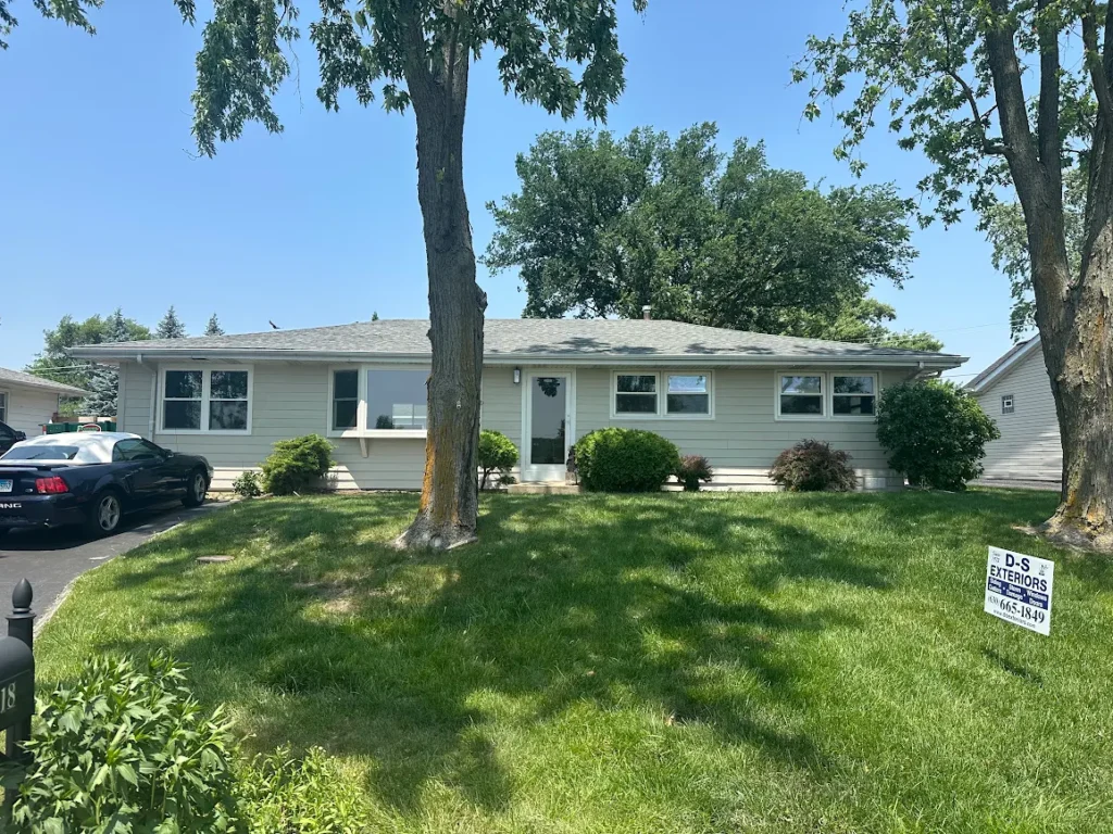 A single-story gray house with a well-kept front lawn, two large trees in front, a black car parked on the driveway, and a yard sign for D-S Exteriors in the grass.