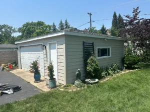 Small gray detached garage with a white paneled door, side entry door, and decorative plants in pots along the front, set in a sunny suburban yard.