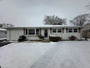 Front view of a ranch-style house with white siding, pink shutters, and a snow-covered yard in winter.