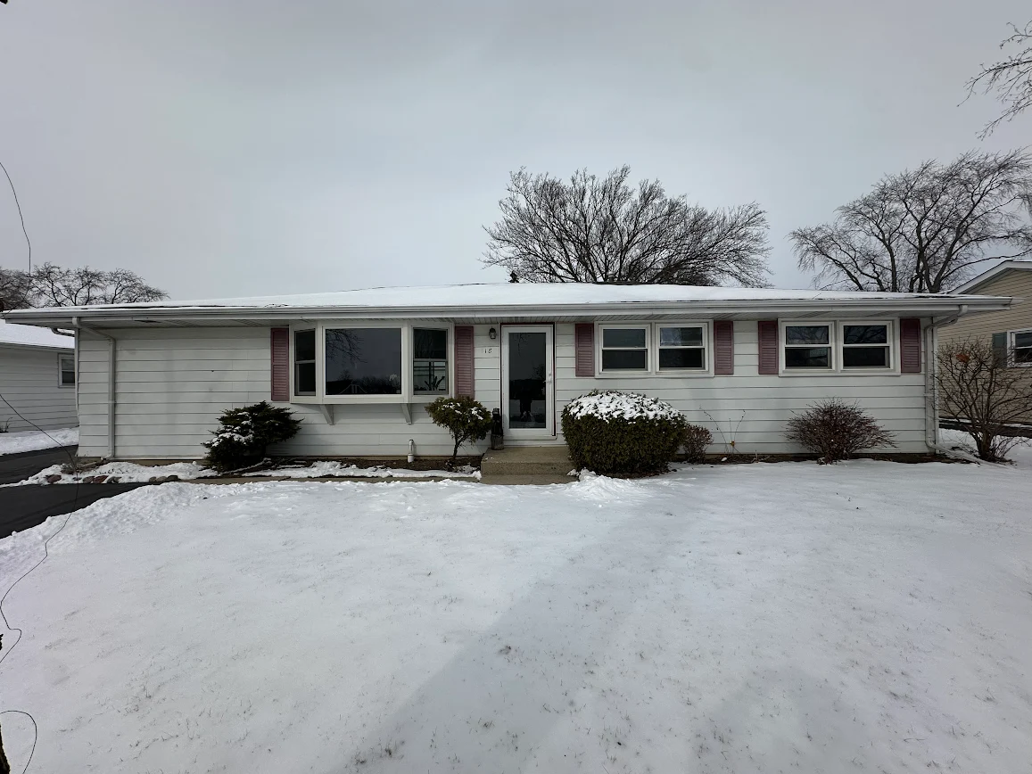 Front view of a ranch-style house with white siding, pink shutters, and a snow-covered yard in winter.