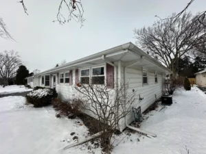 Wintery side view of a single-story white house with snow on the yard and leafless trees nearby, overcast sky above.