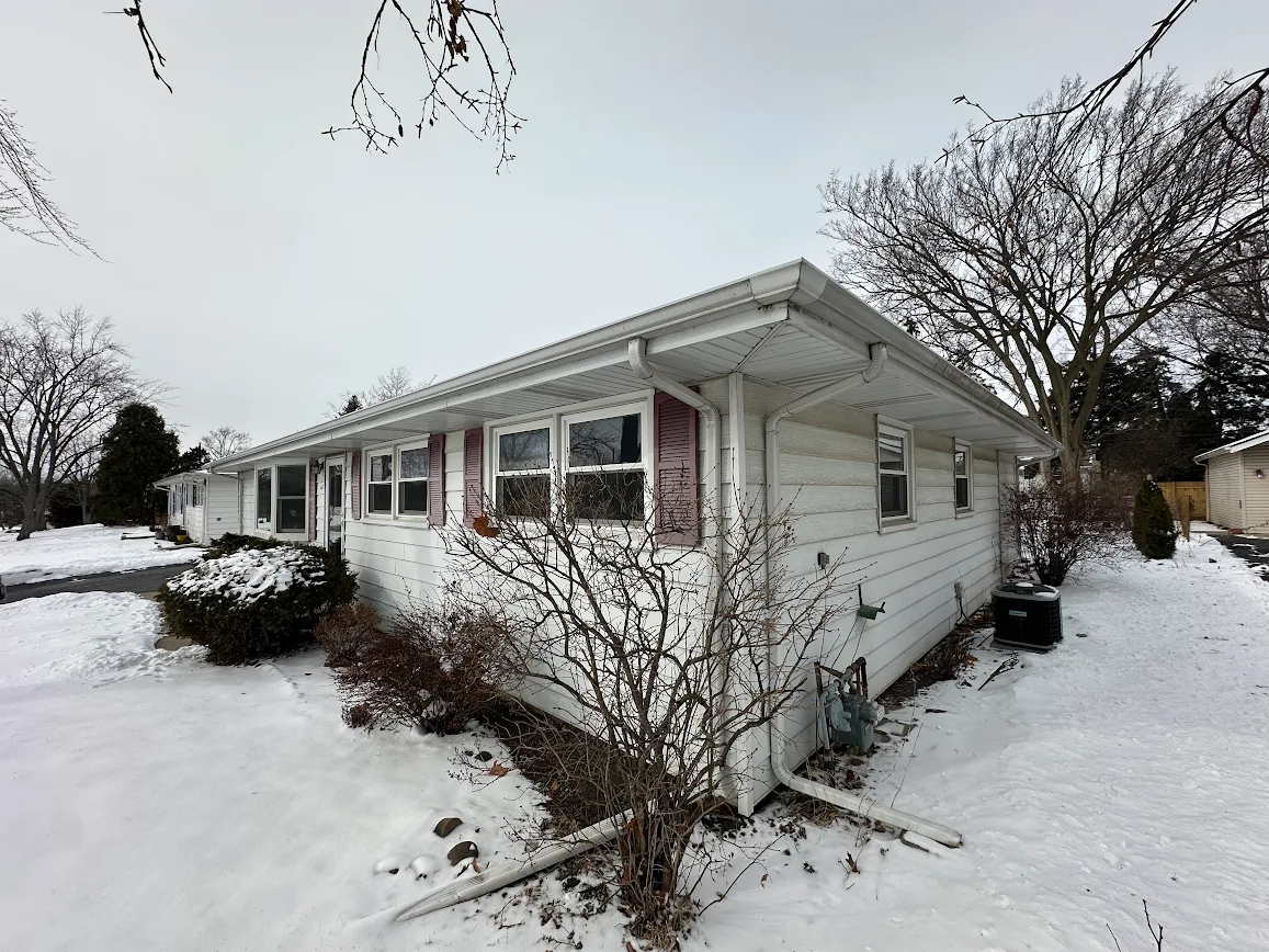 Wintery side view of a single-story white house with snow on the yard and leafless trees nearby, overcast sky above.