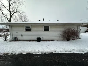 Snow-covered side of a white house with two small windows, an outdoor AC unit, and bare bushes in winter light.