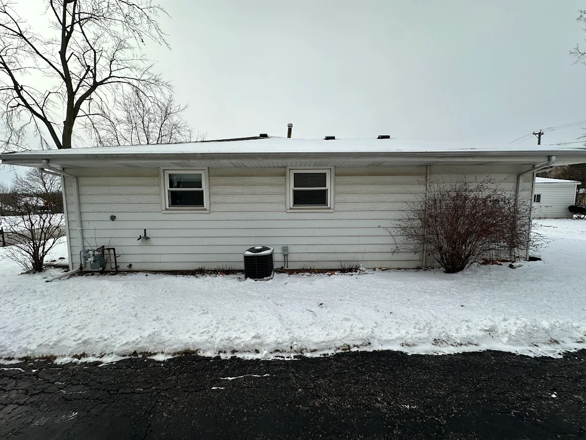 Snow-covered side of a white house with two small windows, an outdoor AC unit, and bare bushes in winter light.