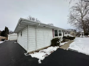 White single-story house with snow along the walkway and driveway, a detached garage in the background, and bare trees lining the street in winter.