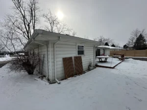 Snowy backyard with a small white siding house; wooden boards lean against the wall and a round table sits on a raised deck.
