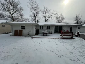 Snowy backyard of a single-story house with a wooden deck, round table, and red chairs under bare trees.