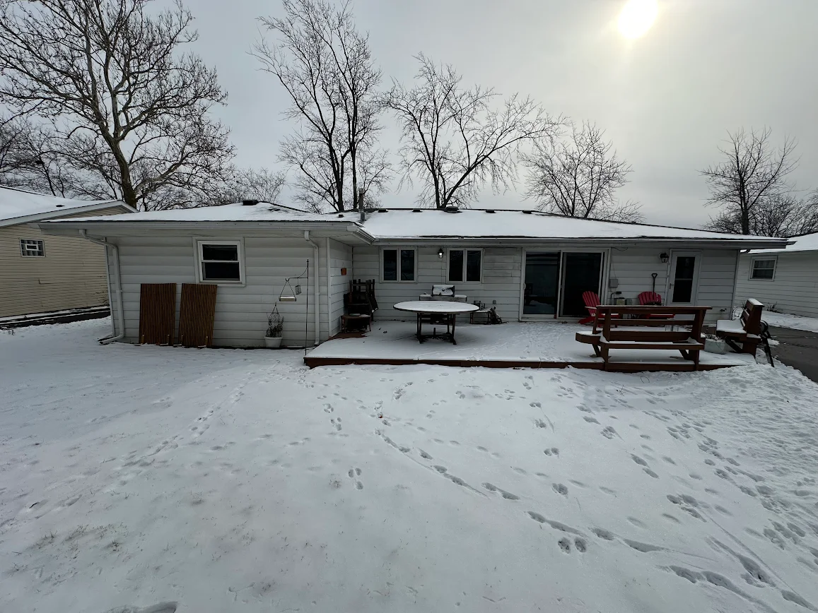 Snowy backyard of a single-story house with a wooden deck, round table, and red chairs under bare trees.