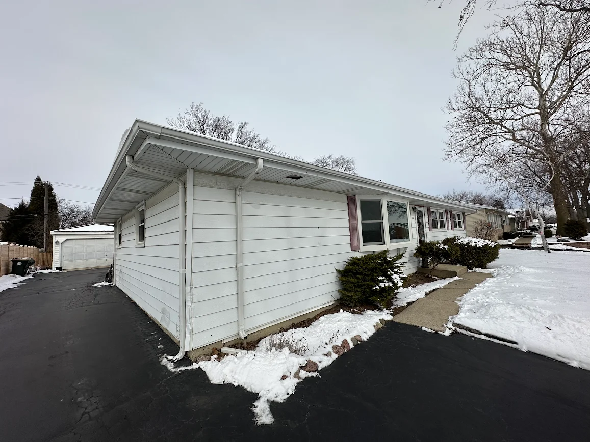 White single-story house with snow along the walkway and driveway, a detached garage in the background, and bare trees lining the street in winter.