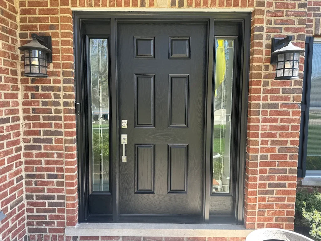 Black paneled front door with narrow glass sidelights flanked by brick walls and two outdoor lanterns.