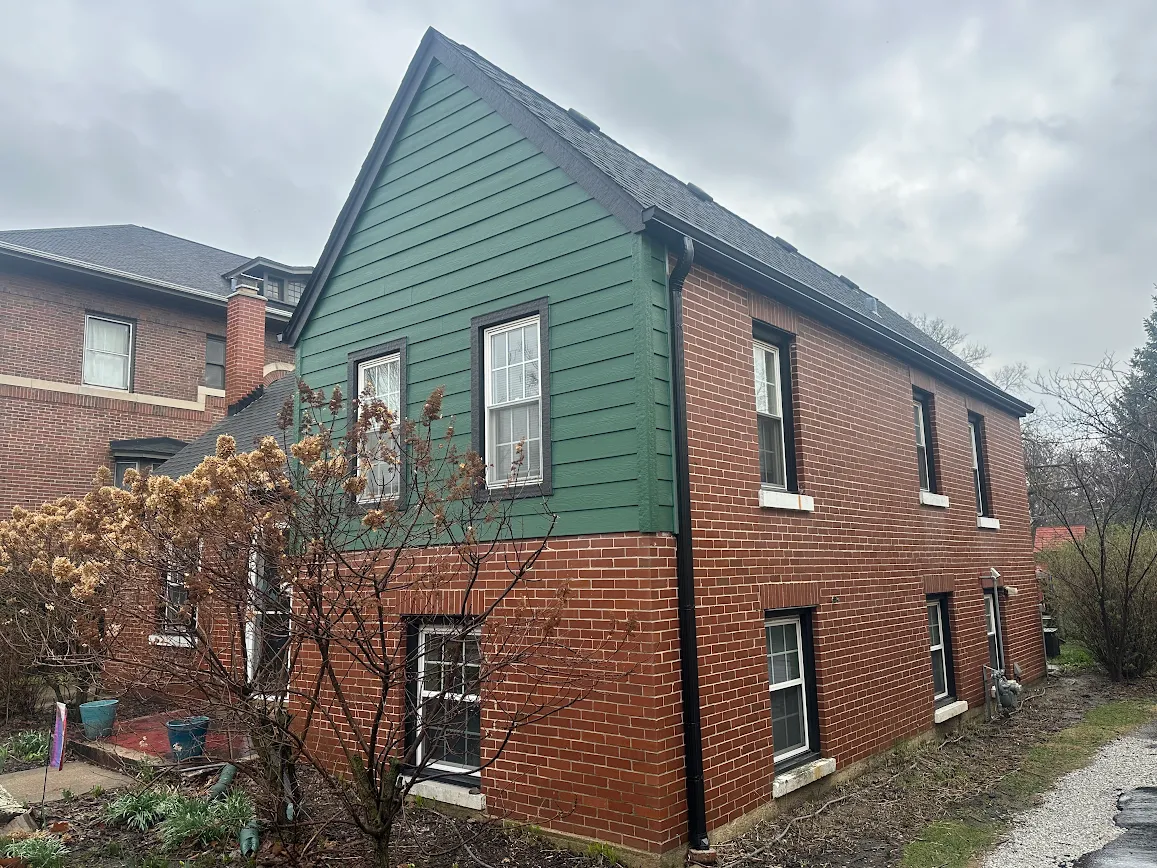 Brick house with green siding on the upper level, white-framed windows, and a flowering tree in a cloudy front yard.
