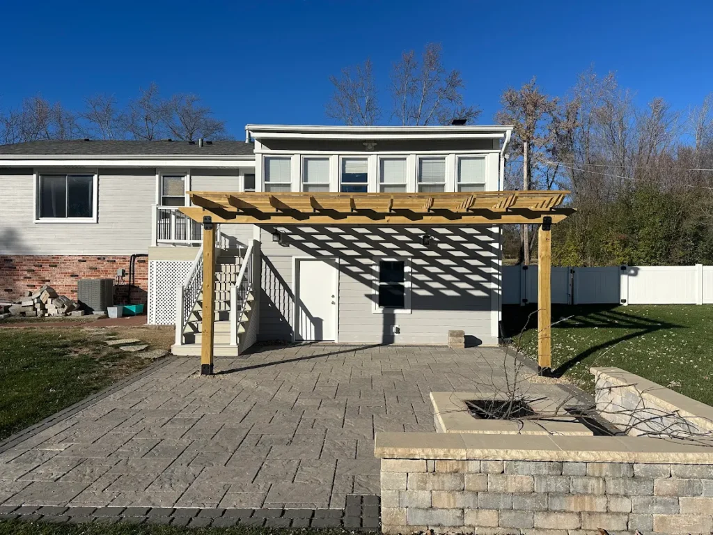 Backyard view of a two-story house with a wooden pergola over a stone-paved patio and a fire pit in a stone planter in the foreground.