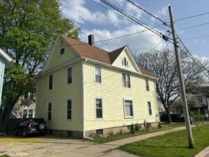 Yellow two-story house with a gabled brown roof on a residential street, with overhead power lines and a parked car nearby.