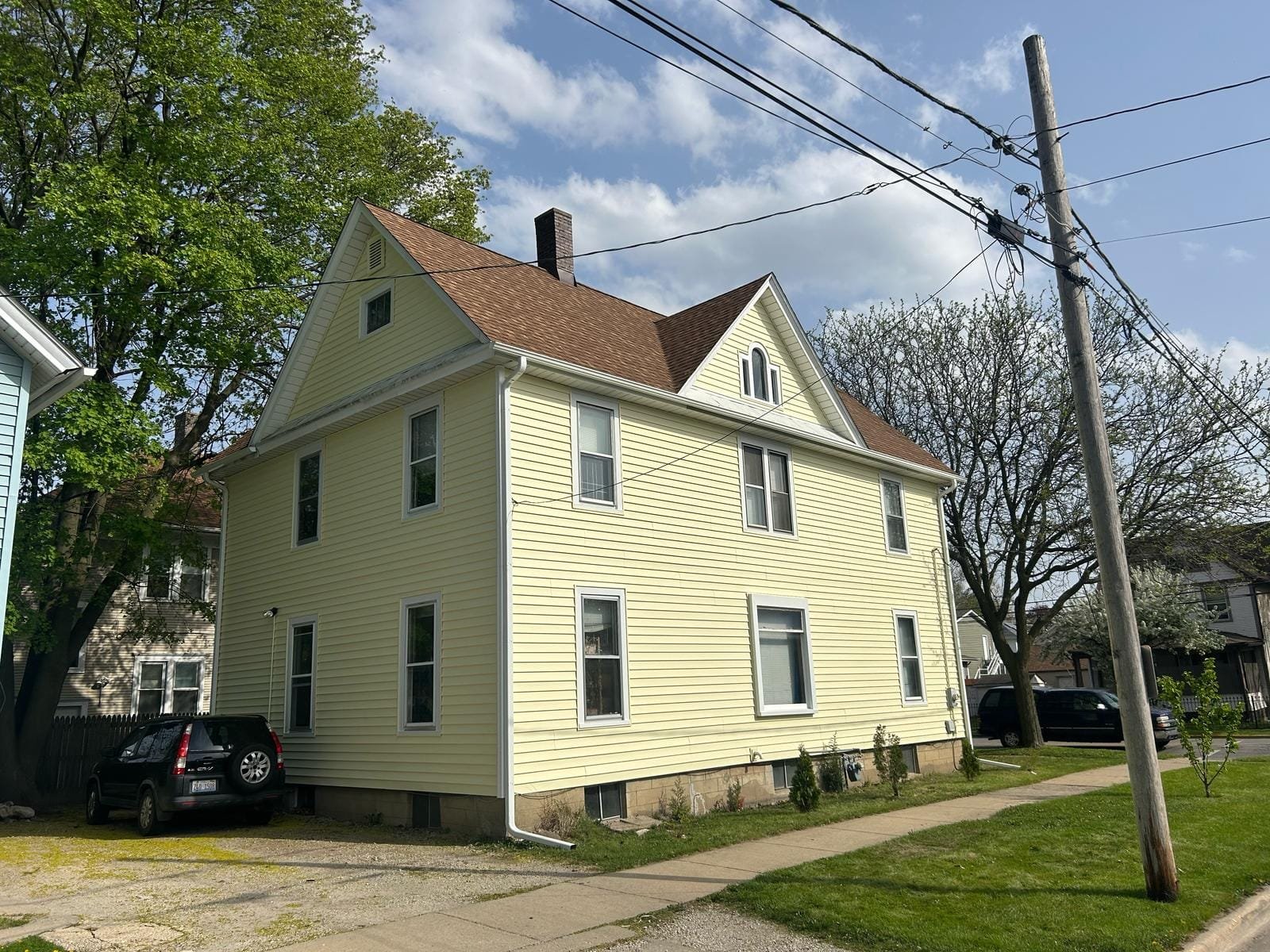 Yellow two-story house with a gabled brown roof on a residential street, with overhead power lines and a parked car nearby.