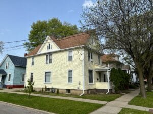 Two-story yellow house with white trim on a sunny street, power lines crossing the frame and trees in bloom across the sky.