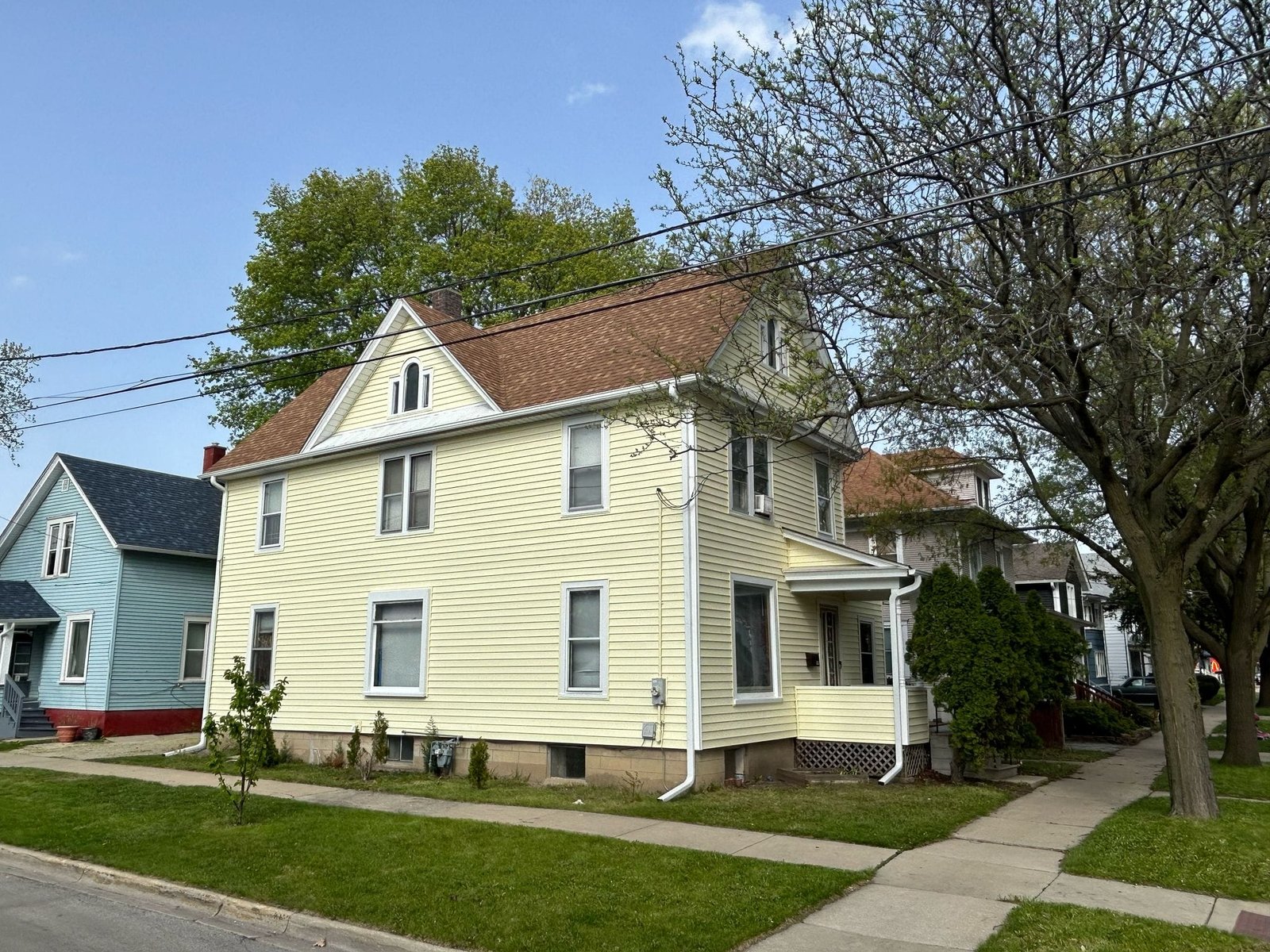 Two-story yellow house with white trim on a sunny street, power lines crossing the frame and trees in bloom across the sky.