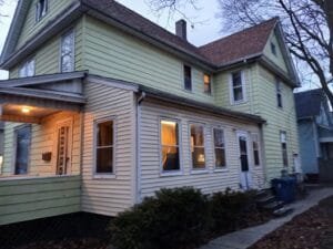 Two-story pale yellow house with a small front porch, lit windows, and leafless trees in the yard at dusk.