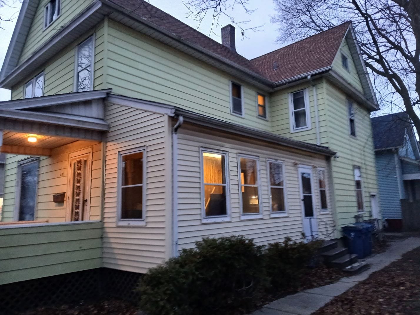 Two-story pale yellow house with a small front porch, lit windows, and leafless trees in the yard at dusk.