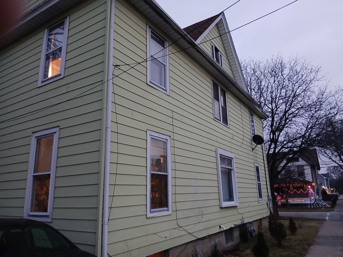 Two-story pale yellow house with multiple white-trimmed windows along a slanted angle, electrical wires overhead, and a tree at the side yard.