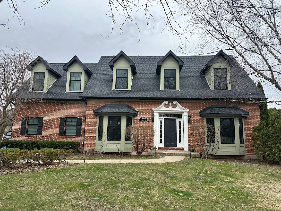 Brick two-story house with a dark roof, three dormers, and green-trimmed bay windows flanking a black front door with white decorative trim.