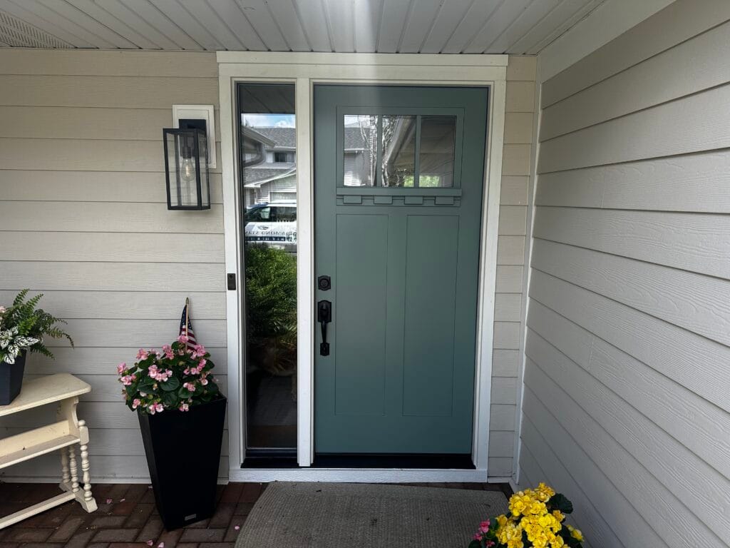 Front porch entry with a green front door, glass sidelights, and a black planter of pink flowers beside a small bench.