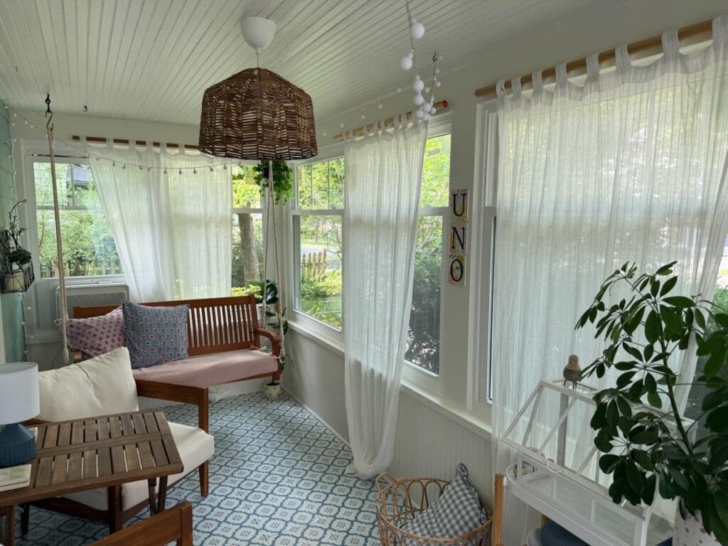 Bright sunroom with sheer white curtains, wooden bench with patterned cushions, and wicker pendant light overlooking a garden.