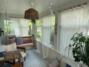 Bright sunroom with sheer white curtains, wooden bench with patterned cushions, and wicker pendant light overlooking a garden.
