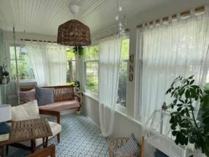 Bright sunroom with sheer white curtains, wooden bench with patterned cushions, and wicker pendant light overlooking a garden.