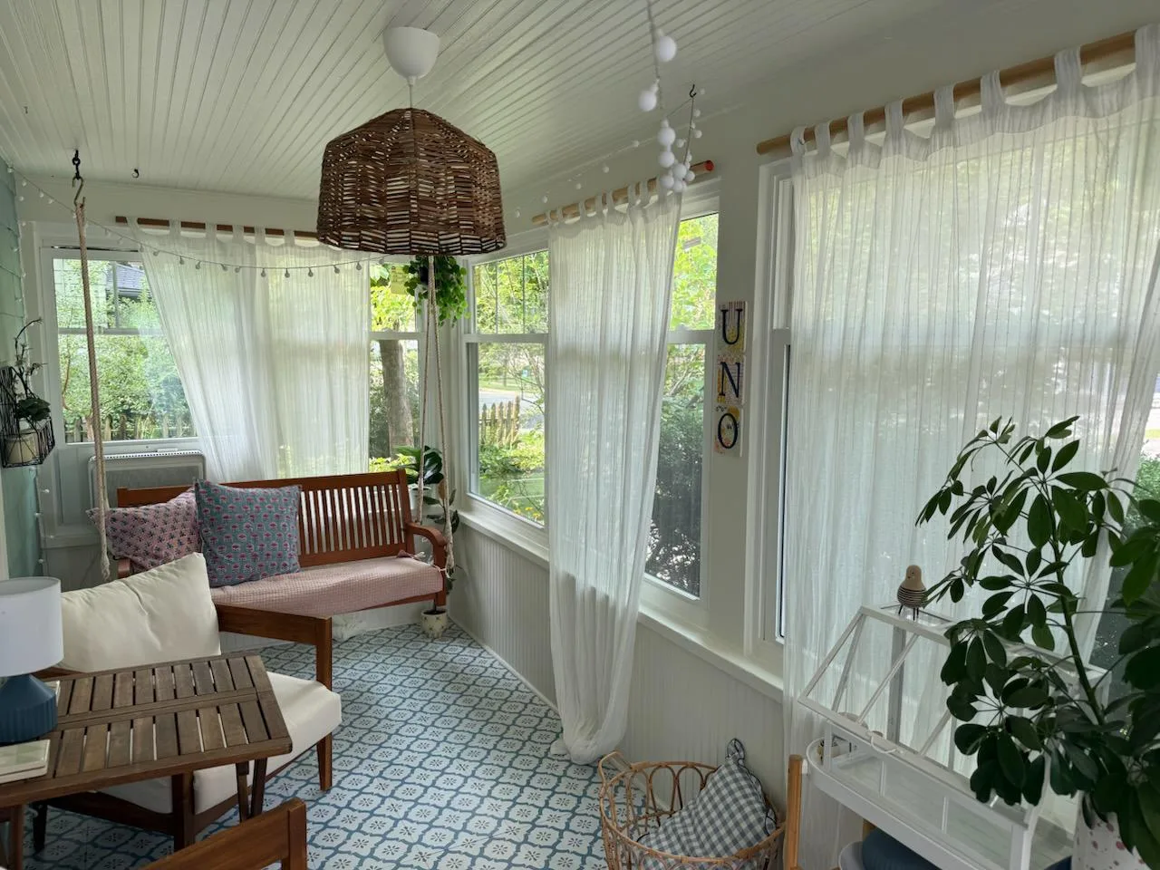 Bright sunroom with sheer white curtains, wooden bench with patterned cushions, and wicker pendant light overlooking a garden.